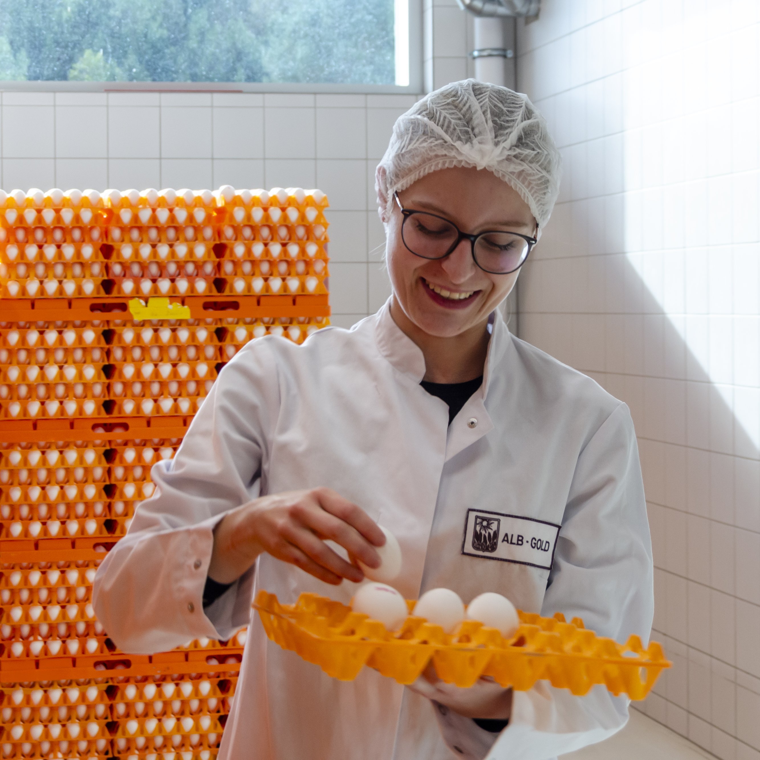 Person in weißem Kittel hält ein gelbes Tablett mit weißen Eiern vor einem Stapel orangefarbener Eierbehälter.