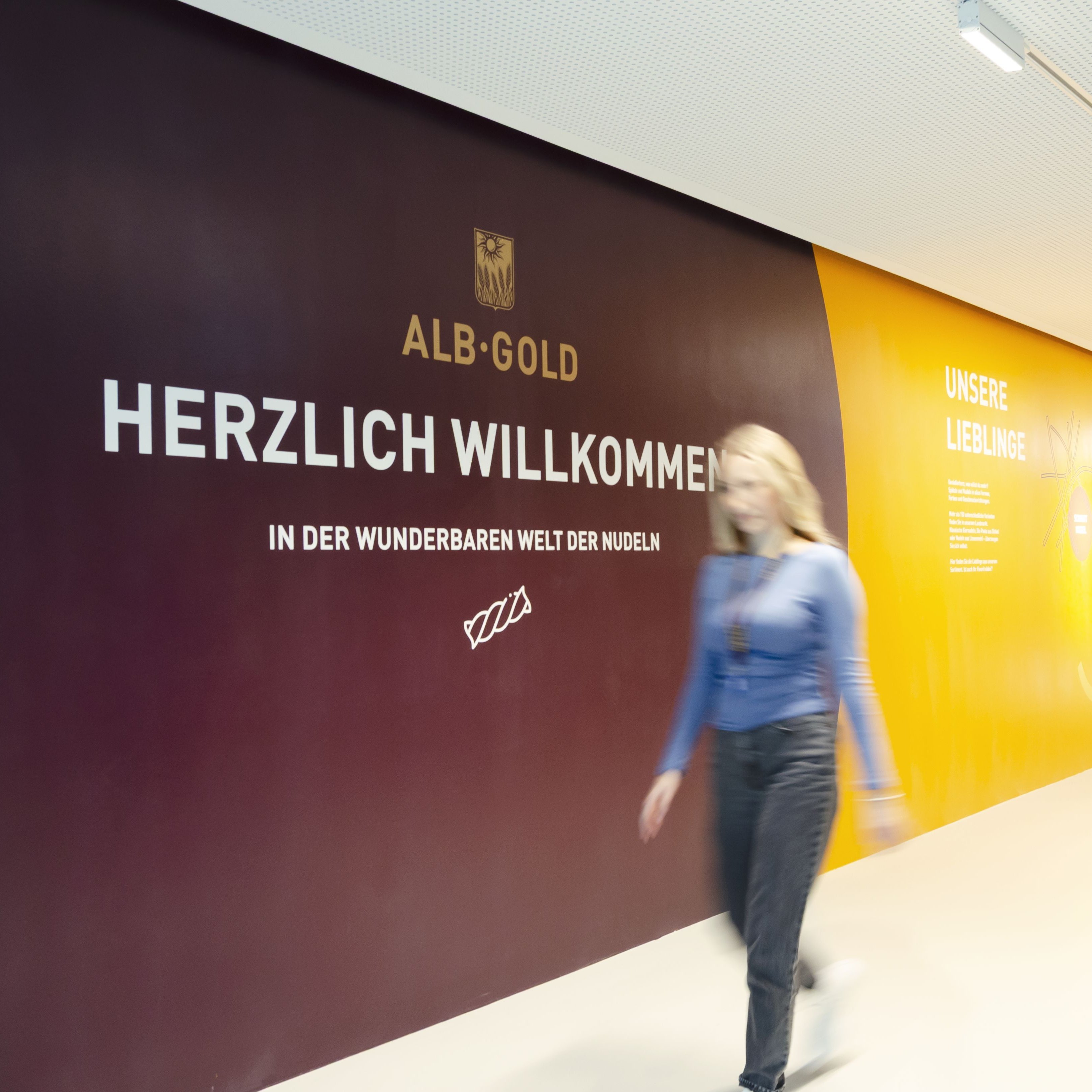 Frau geht an Wand mit Text 'Herzlich Willkommen' und 'Alb-Gold' Logo in einem hellen Flur entlang
