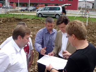 Vier Männer besprechen Pläne auf einer Baustelle mit gegrabenem Boden und parkenden Autos im Hintergrund