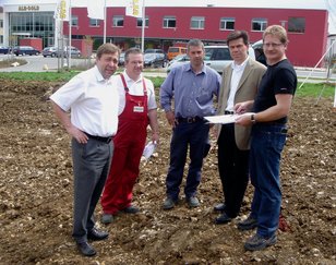 Fünf Männer stehen auf einer Baustelle, einer hält Pläne, im Hintergrund ein rotes Gebäude