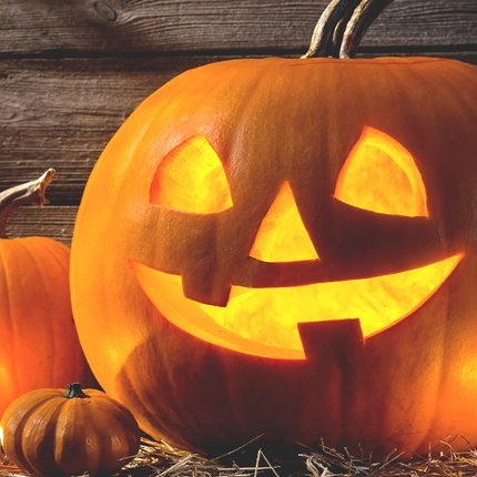 Leuchtender Halloween-Kürbis mit ausgeschnittenen Augen, Nase und lachendem Mund, umgeben von weiteren Kürbissen auf Holzboden vor Holzwand.