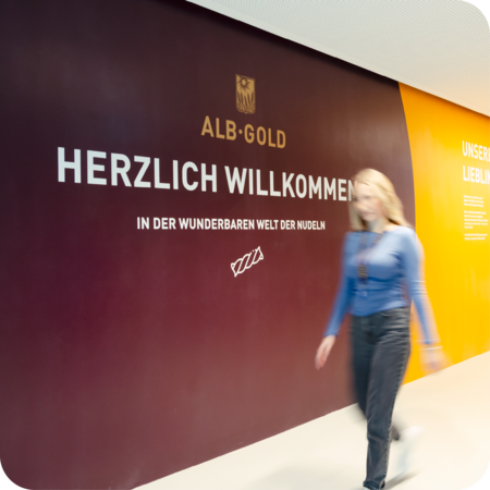 Frau geht an einer Wand mit der Aufschrift 'ALB-GOLD HERZLICH WILLKOMMEN IN DER NUDELWELT' vorbei.