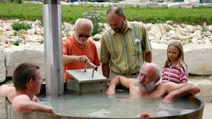 Mehrere Personen bei der Einweihung eines runden Wasserbeckens im Rahmen des Sommermarktes, zwei Männer bedienen eine Vorrichtung, drei Personen sind im Wasser, darunter ein älterer Mann und ein Mädchen