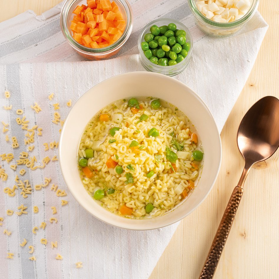 Schale mit Buchstabennudelsuppe mit Erbsen und Karotten, daneben drei Gläser mit gewürfelten Karotten, Erbsen und Sellerie, brauner Löffel