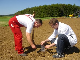 Zwei Männer untersuchen den Boden auf einem Acker, einer trägt rote Arbeitshosen und hält einen Spaten, der andere kniet und hält eine Bodenprobe