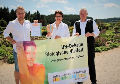 Drei Personen halten eine Urkunde, eine kleine Trophäe und ein Banner mit der Aufschrift 'UN-Dekade Biologische Vielfalt Ausgezeichnetes Projekt' bei der Auszeichnung eines Gartens im Freien
