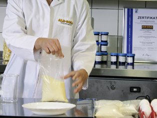 Person in weißem Laborkittel hält einen durchsichtigen Beutel mit gelblicher Substanz über einem weißen Teller auf einem Labortisch.