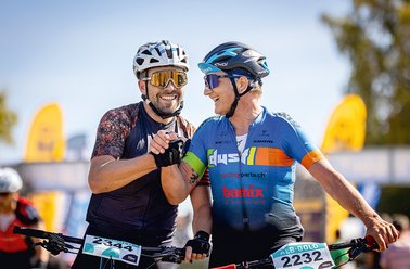 Zwei Radfahrer in Helmen und Sportkleidung geben sich lachend die Hand bei einem Radrennen.