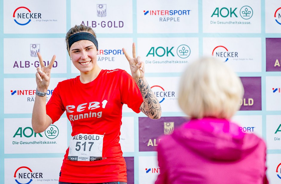 Teilnehmerin eines Laufs in rotem Shirt mit Startnummer 517 zeigt zwei Fingerzeichen und lächelt in die Kamera, im Hintergrund Sponsorenbanner
