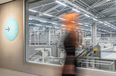 Eine vorbeilaufende Frau an einem großes Fenster mit Sicht auf die Nudelproduktion.