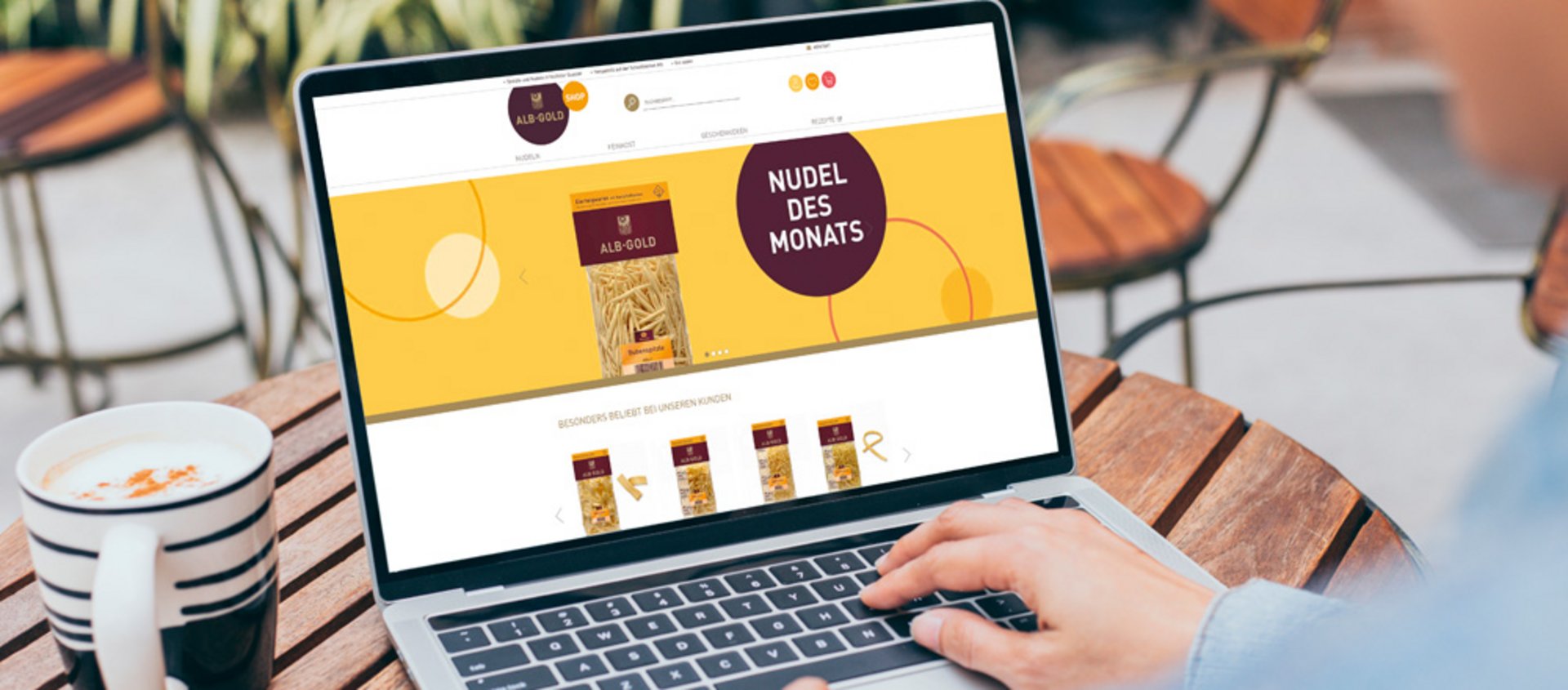 Laptop zeigt Webseite mit 'Nudel des Monats' und verschiedenen Pasta-Produkten auf einem Holztisch mit Kaffeetasse