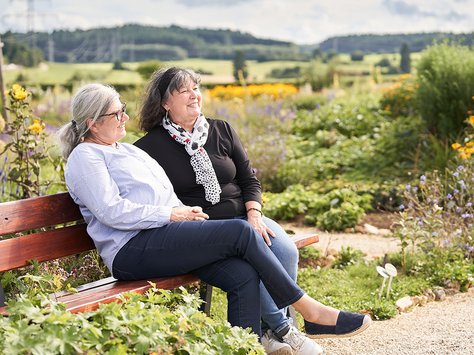 Zwei ältere Frauen sitzen entspannt auf einer Parkbank in einem blühenden Garten mit Blick auf eine ländliche Landschaft.