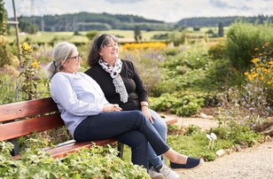 Zwei ältere Frauen sitzen auf einer Bank in einem blühendem Garten