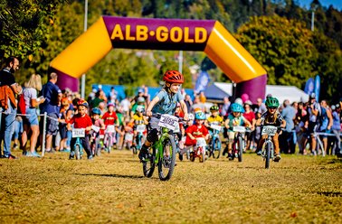 Kinder fahren Fahrrad bei einem Rennen unter einem gelb-lila Startbogen mit der Aufschrift 'ALB-GOLD'