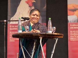 Vanda Shiva am Podium mit Wasserflaschen und Gläsern.