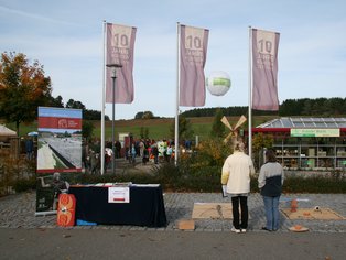 Zwei Personen stehen vor einem Informationsstand mit drei Fahnen, im Hintergrund Bäume und ein Gebäude