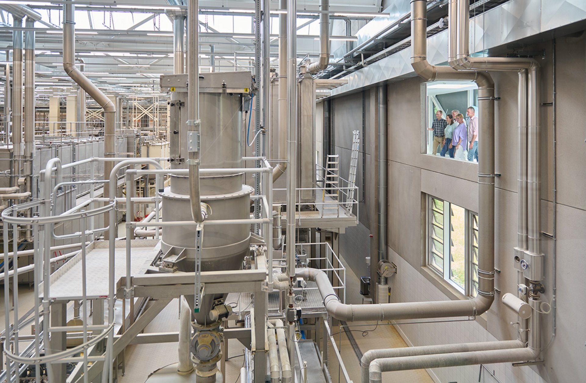 Industrielle Anlage mit zahlreichen Rohren, Ventilen und Metallstrukturen in einer großen Halle mit Fenster, durch das Menschen zu sehen sind