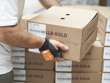 Person scanning barcode on a brown cardboard box labeled 'ALB-GOLD' in a warehouse