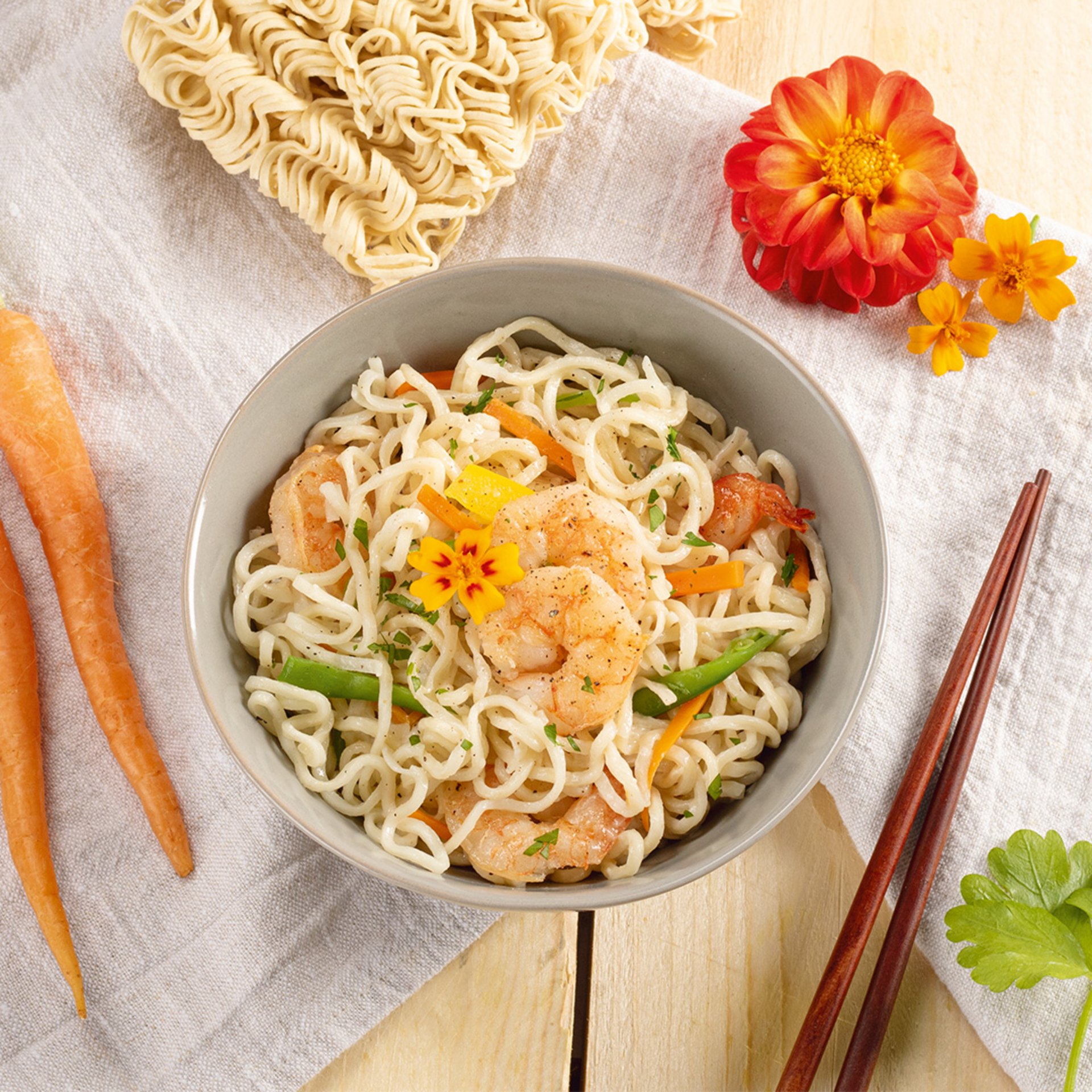 Bowl of mie noodles with shrimp, vegetables, and garnished with a flower, accompanied by chopsticks and uncooked noodles on the side