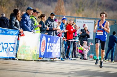 Läufer in blauer Sportkleidung läuft bei einem Straßenrennen, Zuschauer stehen hinter Absperrungen und feuern an
