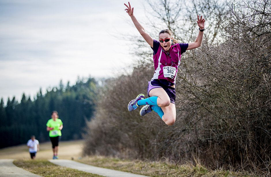 Junge Frau in Sportkleidung springt mit erhobenen Armen auf einem Waldweg während eines Laufs