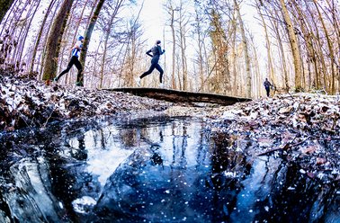Zwei Personen springen über einen Bach in einem kahlen Wald im Winter