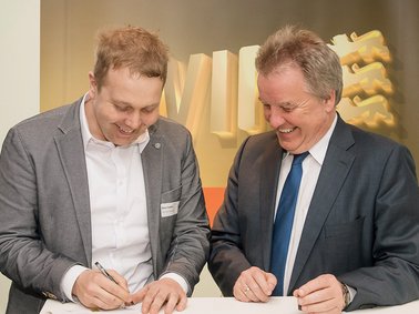 Two men in business attire signing documents together at a table, both smiling and focused.