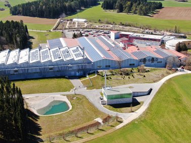 Aerial view of a large industrial facility surrounded by green fields and trees, with multiple connected buildings and a parking area.