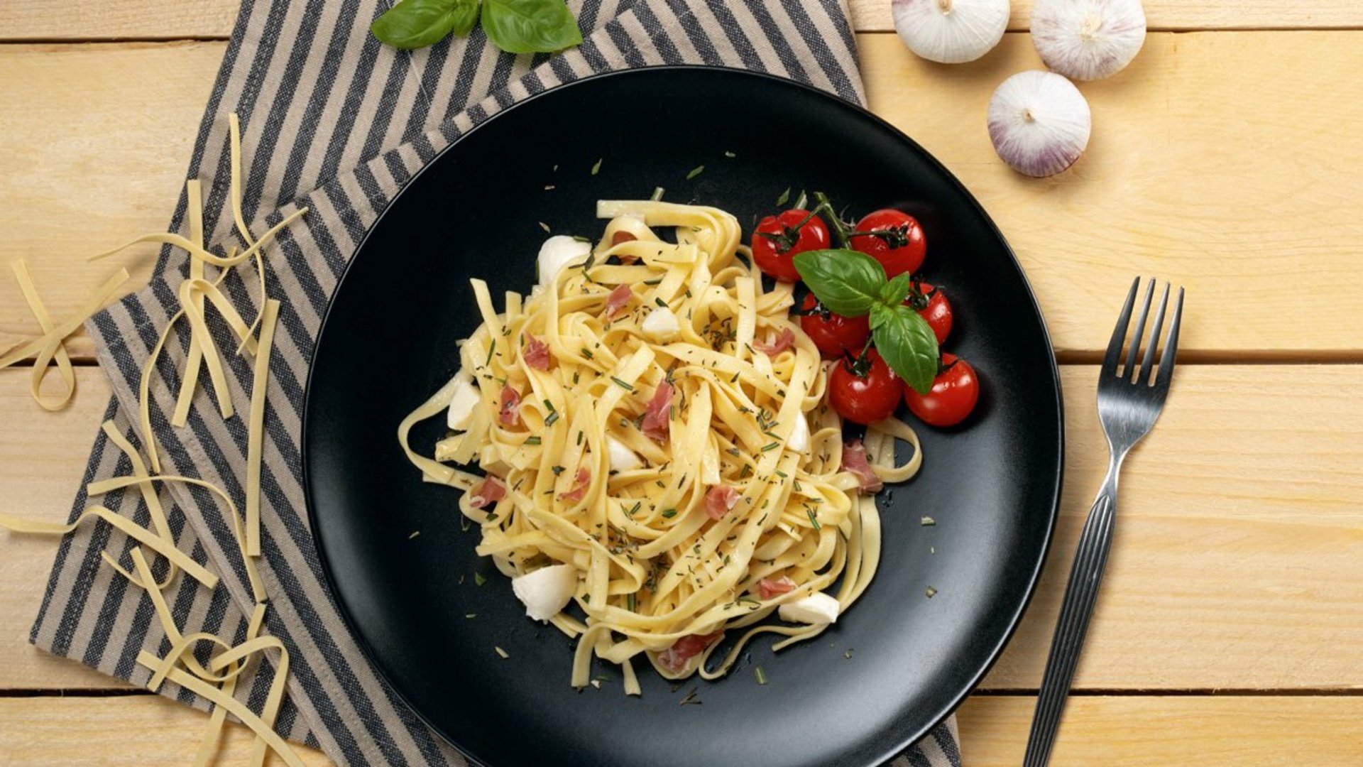 Schwarzer Teller mit Spaghetti Carbonara, Kirschtomaten und Petersilie auf Holztisch mit Gabel und gestreiftem Tuch