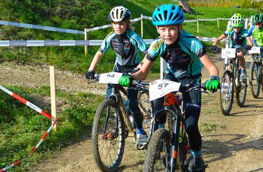 Kinder fahren Mountainbikes auf einem markierten Waldweg, vorne ein Junge mit blauem Helm und Nummer 57 am Fahrrad