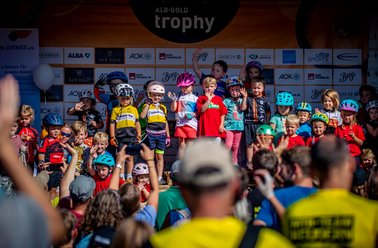 Kinder in bunten Fahrradhelmen und Trikots stehen auf einer Bühne bei einer Siegerehrung, Publikum fotografiert und filmt sie