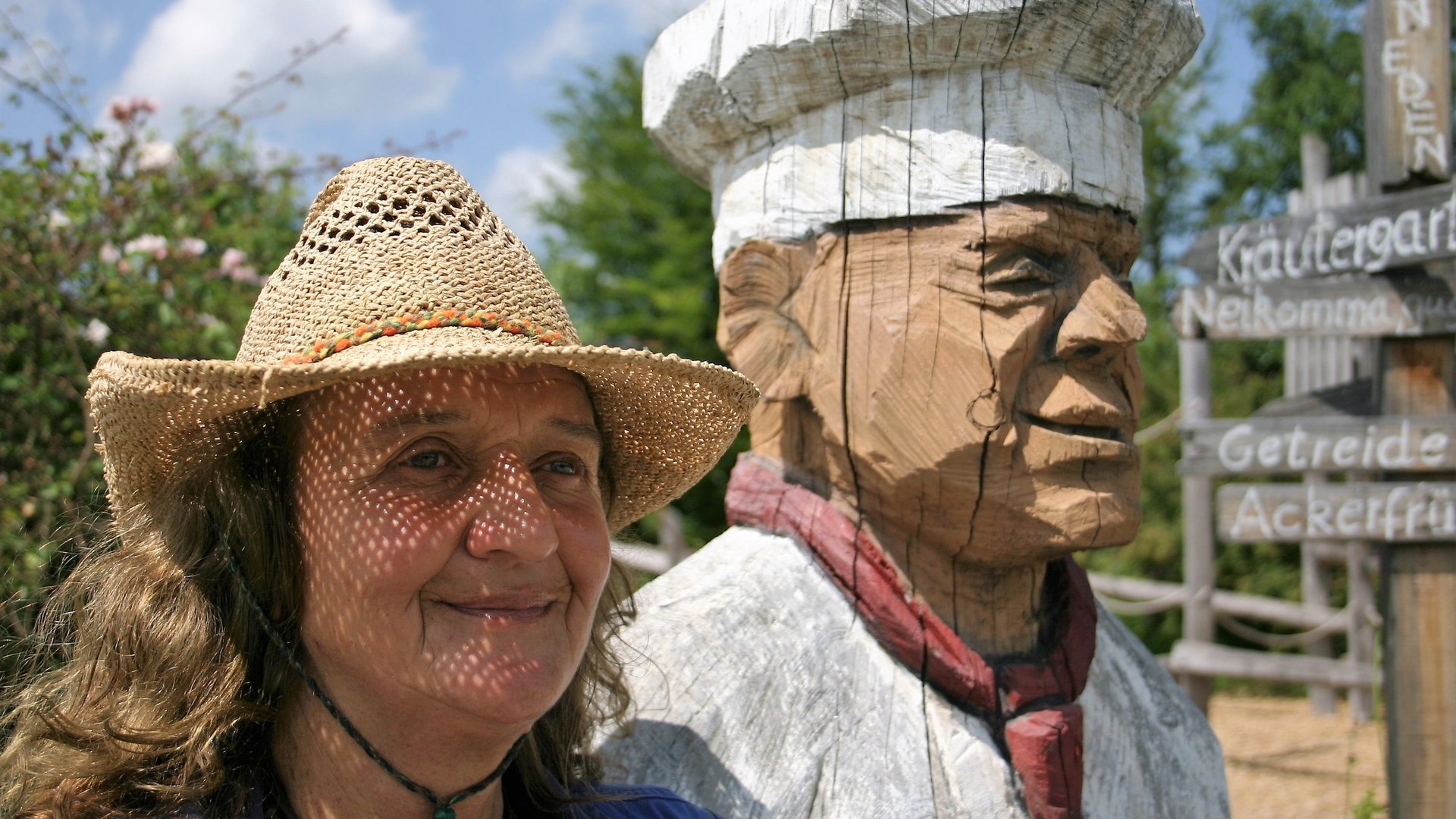 Eine Frau mit einem Strohhut steht neben einer Holzfigur eines Kochs.