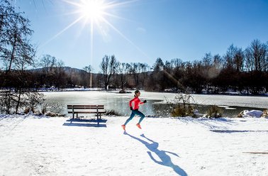 Person in roter Jacke läuft im Schnee entlang eines zugefrorenen Sees bei sonnigem Winterwetter