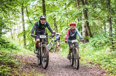 Zwei Mountainbiker fahren auf einem Pfad durch den Wald.