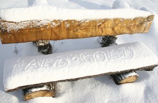 Eine Sitzbank ist mit Schnee bedeckt, es wurden die Worte ALB-GOLD auf die Bank geschrieben und befindet sich in einer Schneelandschaft