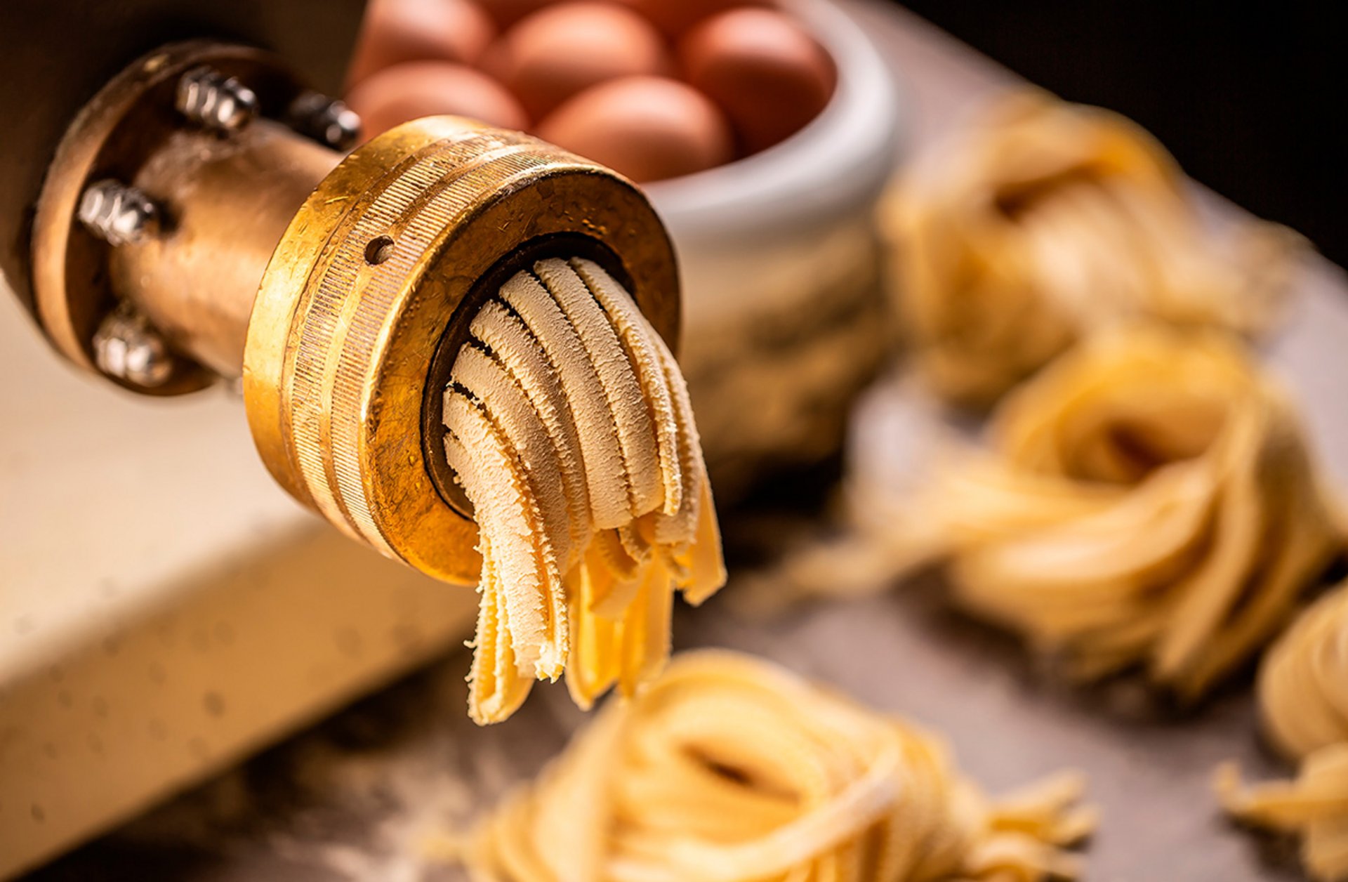 Fresh pasta dough being extruded through a wooden pasta maker with eggs and pasta nests in the background