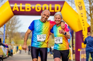 Zwei ältere Läufer in bunten Shirts mit Startnummern 479 und 478 lächeln und umarmen sich unter einem gelben Zielbogen mit der Aufschrift 'ALB-GOLD'