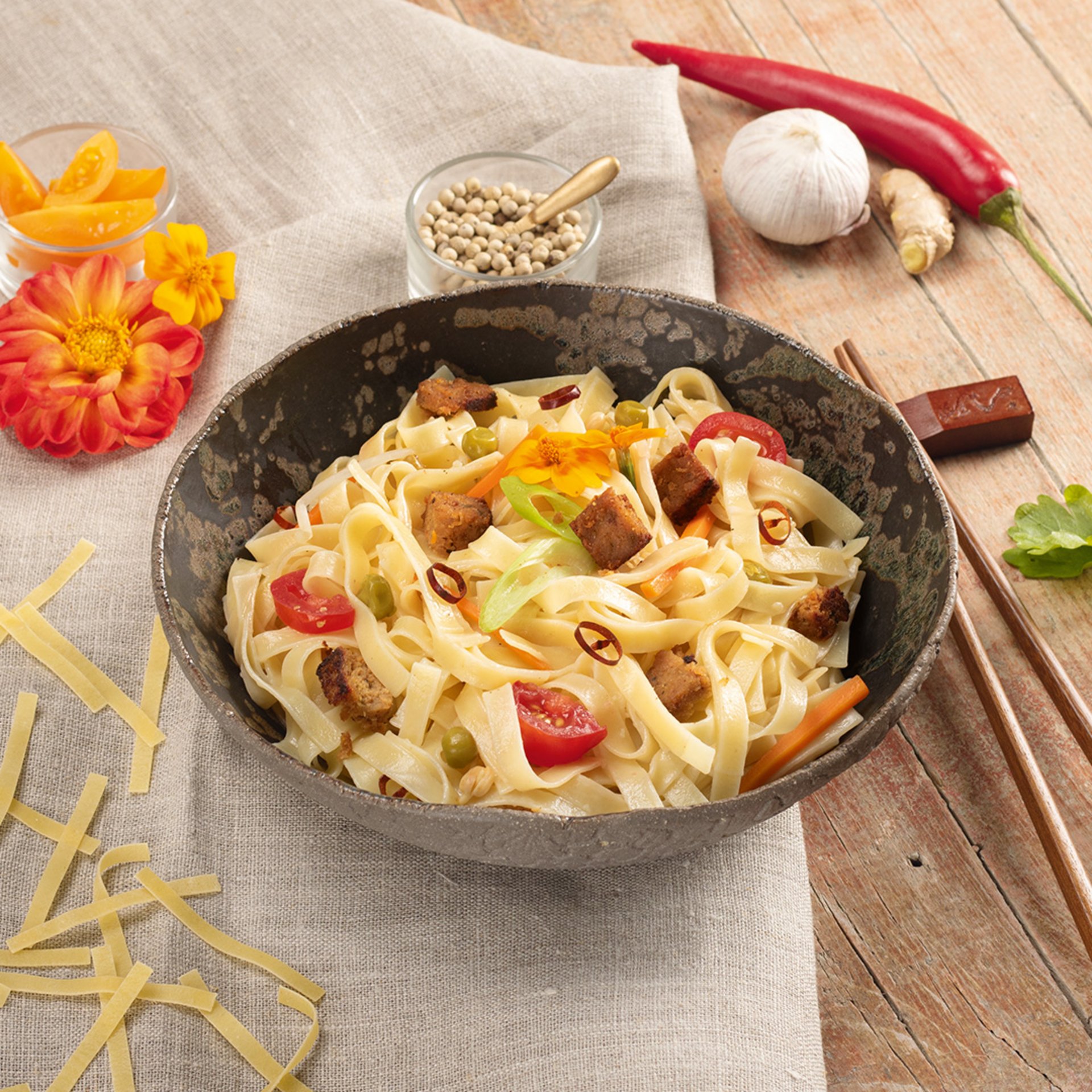 Wok noodles with vegetables and tofu in a black bowl, accompanied by chopsticks and ingredients like garlic and chili on a light cloth