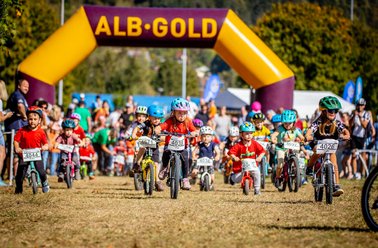 Kinder starten ein Fahrradrennen unter einem gelben Startbogen mit der Aufschrift 'ALB-GOLD' auf einem Feldweg