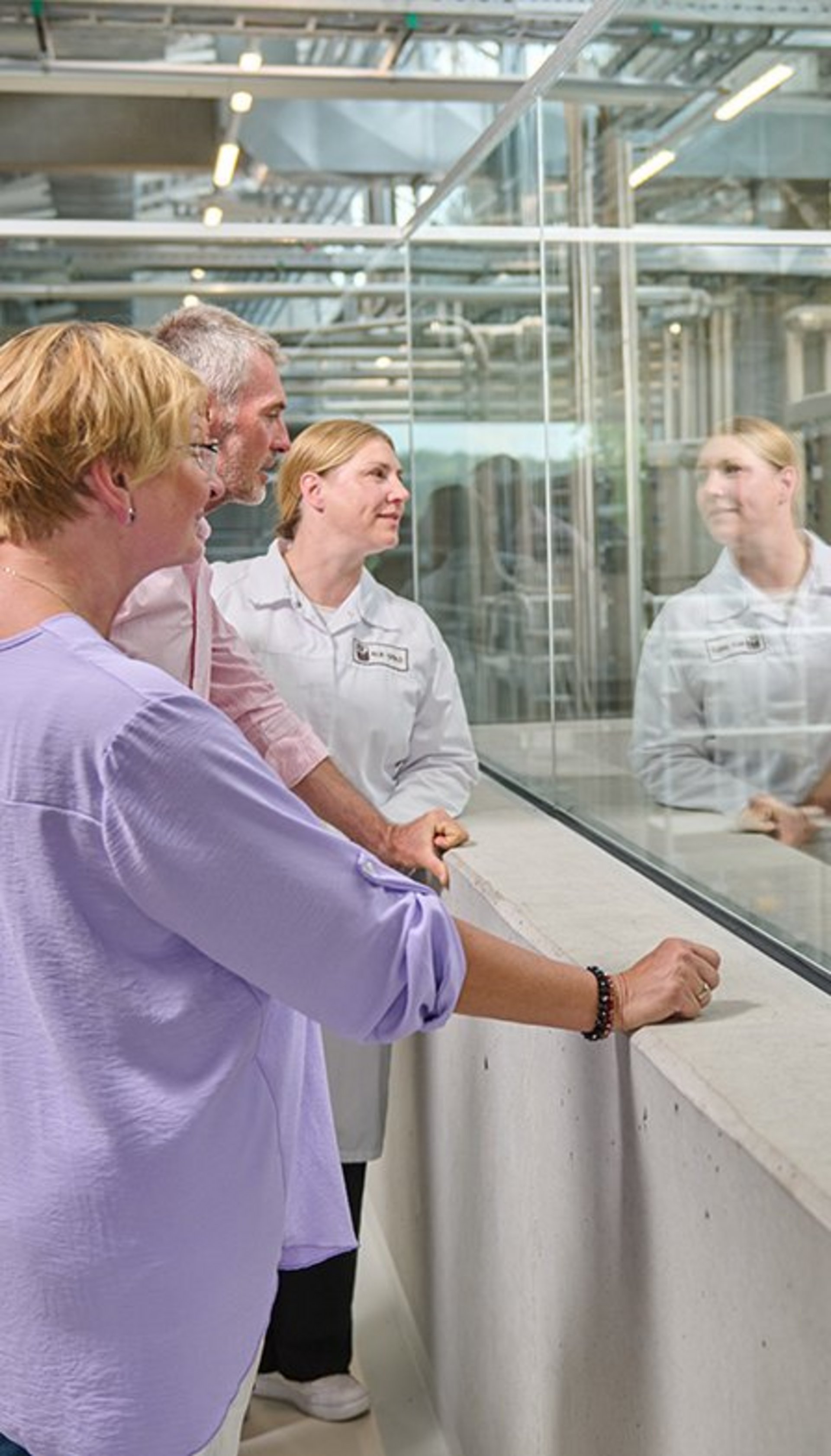Drei Personen in einem Labor oder einer Produktionsanlage, zwei tragen weiße Laborkittel, sie unterhalten sich durch eine Glasscheibe
