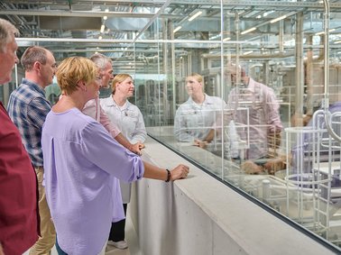 Group of five people observing a factory production line through a glass barrier