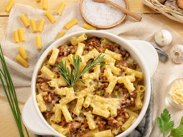 Baked rigatoni pasta with ground meat and melted cheese in a white ceramic dish, garnished with rosemary on a wooden table with garlic, cheese, and uncooked pasta nearby