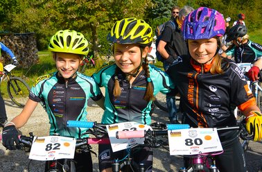 Drei Kinder mit Fahrradhelmen und Trikots stehen nebeneinander mit ihren Mountainbikes bei einem Rennen, Nummernschilder 82, 2 und 80 sichtbar