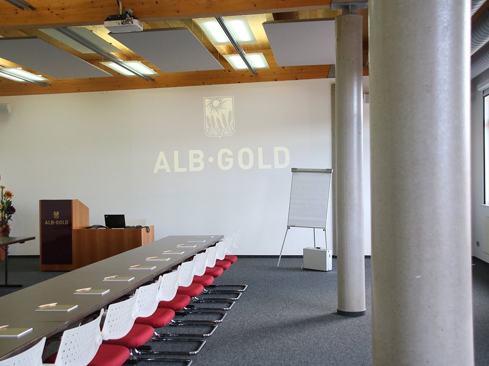 Konferenzraum mit langem Tisch, weißen Stühlen mit roten Sitzflächen, Flipchart und Projektionswand mit Schriftzug 'ALB-GOLD'