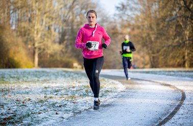 Frau in pinker Jacke läuft auf einem verschneiten Weg im Park, ein weiterer Läufer folgt ihr