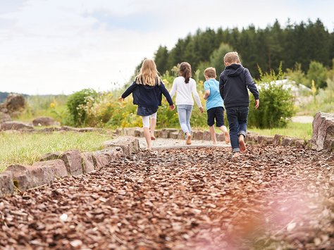 Vier Kinder laufen auf einem mit Holzschnitzeln bedeckten Weg in einer grünen Parklandschaft mit Bäumen im Hintergrund