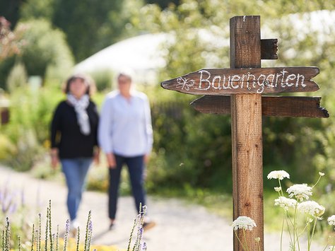 Holzschild mit der Aufschrift 'Bauerngarten' zeigt den Weg in einen Garten, zwei Personen gehen im Hintergrund auf einem Weg