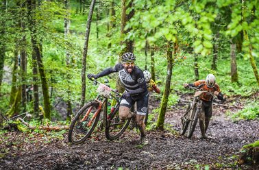 Zwei Mountainbiker kämpfen sich durch einen schlammigen Waldweg.