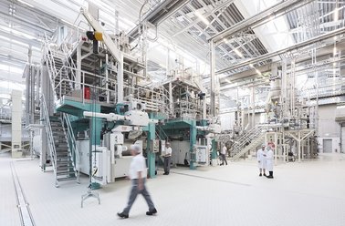 Große industrielle Produktionsanlage mit komplexen Maschinen und zwei Personen in Schutzkleidung in hellem, sauberem Raum
