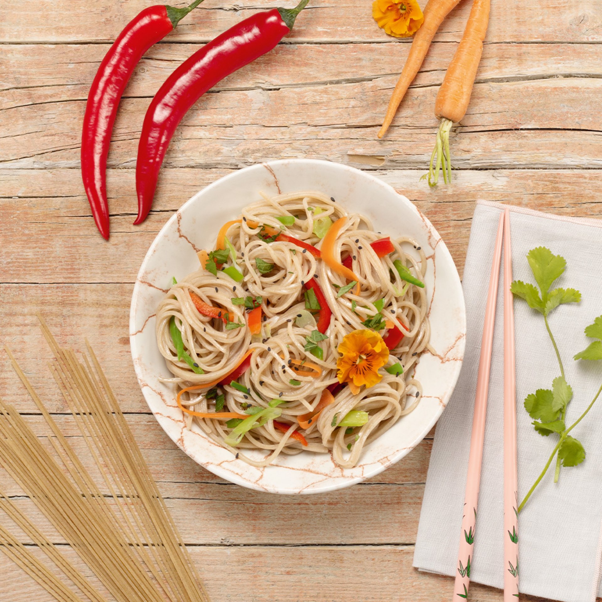 Bowl of soba noodles mixed with sliced vegetables on a wooden table, accompanied by chopsticks and two red chili peppers
